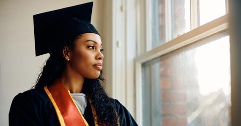 Pensive African American woman in graduation gown looks through window.