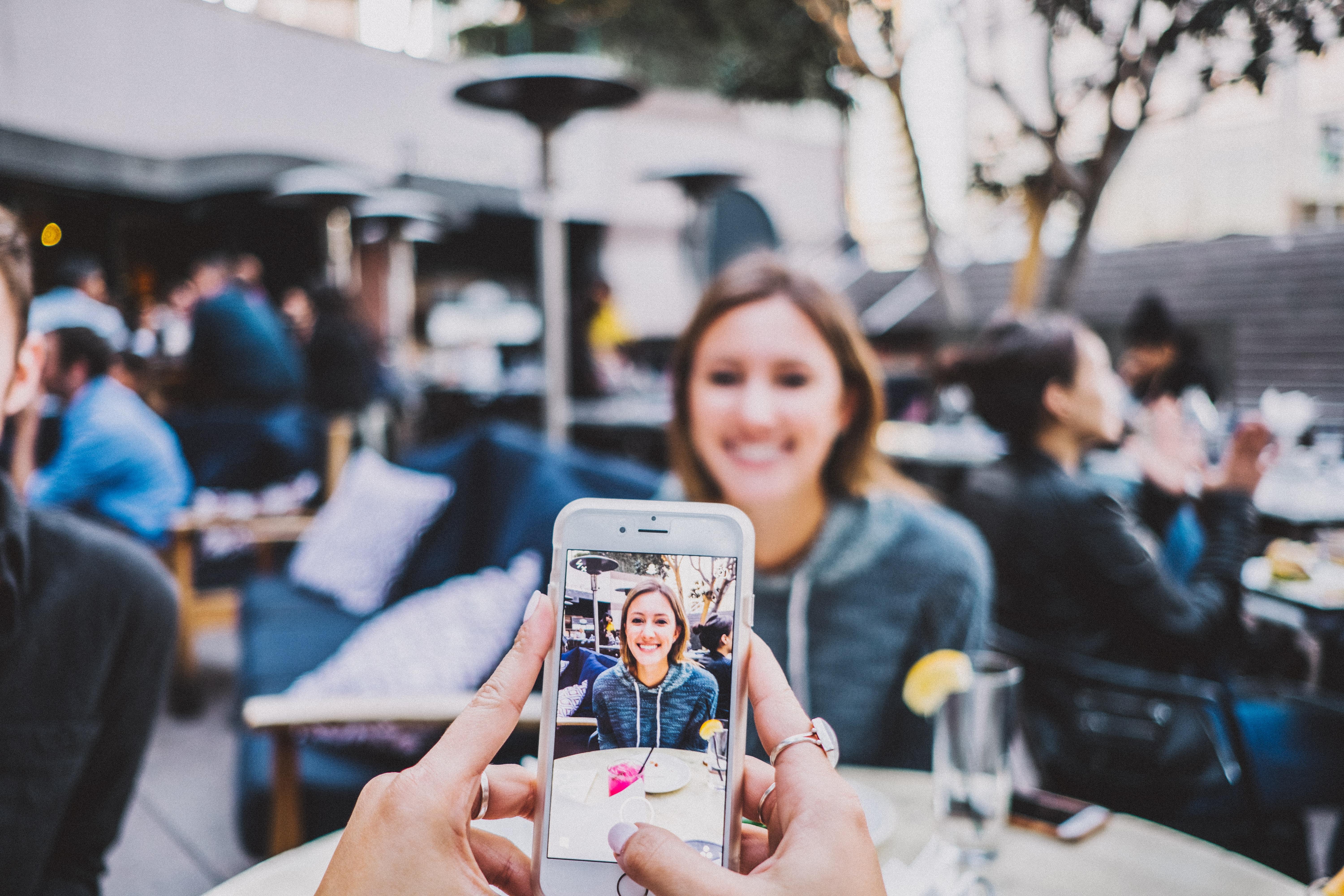 A person taking a photo of a smiling woman sitting at a table with an iPhone