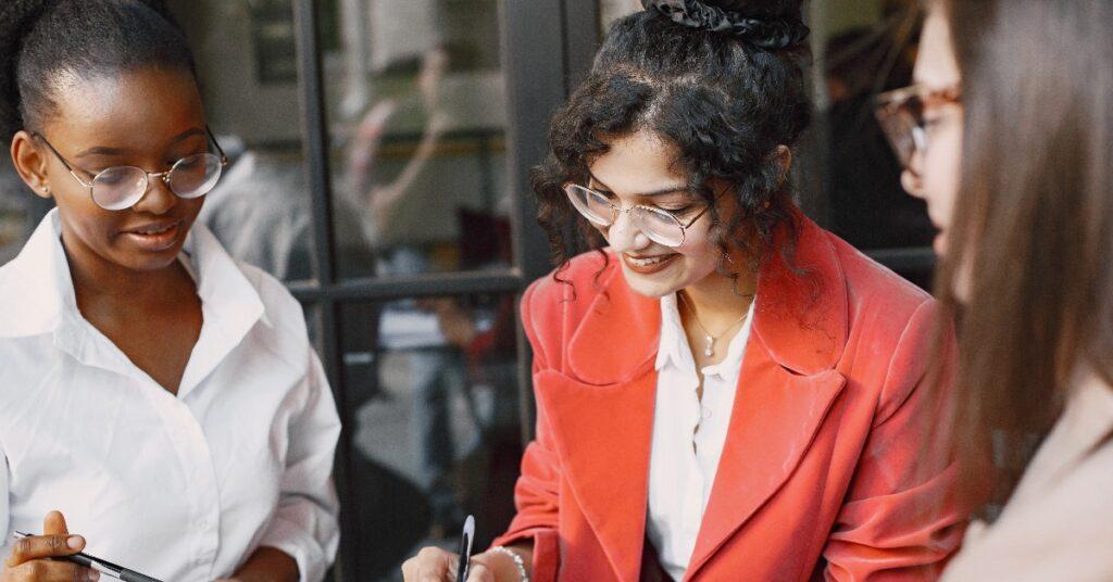 three women sit at a table together looking downwards with pens in their hands