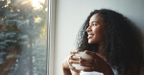 Morning routine. Portrait of happy charming young mixed race female with wavy hair enjoying summer view through window, drinking good coffee, sitting on windowsill and smiling. Beautiful daydreamer