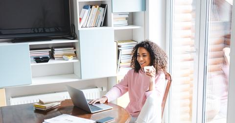 happy african american freelancer holding cup while using laptop at home