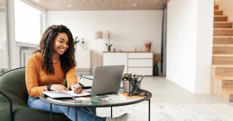 Smiling woman sitting at table, using computer writing in notebook