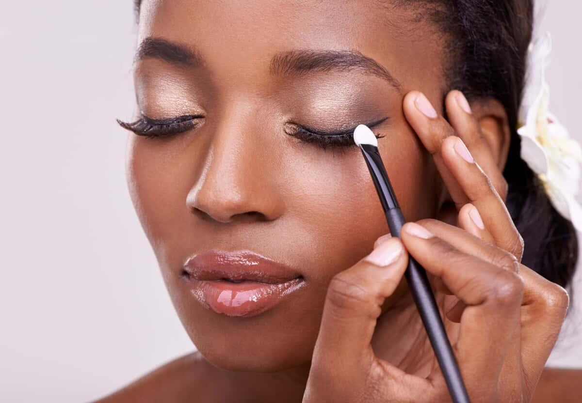 Black woman, face and brush for eyeshadow with beauty, makeup and lashes on grey background. Skin glow, hands and apply cosmetic product with tools for cosmetology, shimmer or glitter with shine