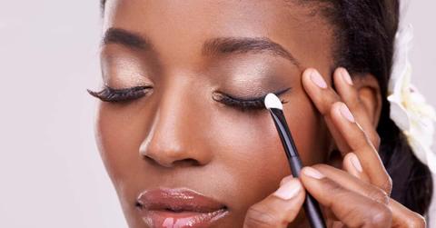 Black woman, face and brush for eyeshadow with beauty, makeup and lashes on grey background. Skin glow, hands and apply cosmetic product with tools for cosmetology, shimmer or glitter with shine