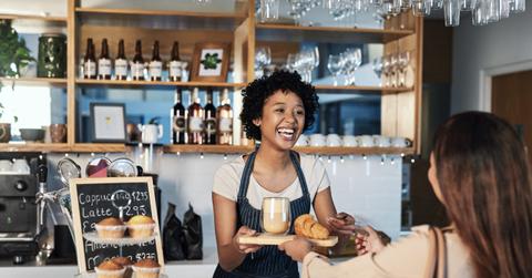 Happy woman, barista and serving customer at cafe for service, payment or order on counter at coffee shop. African person, waitress or employee in small business restaurant helping client at checkout