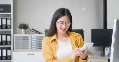 A young Asian woman works at her desk in an office, surrounded by paper files, financial charts, and devices. She focuses on HR, legal consulting, and compliance while managing time effectively.
