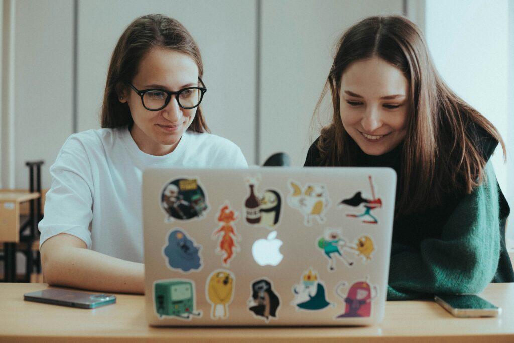 Two women viewing a screen on a sticker-decorated laptop.