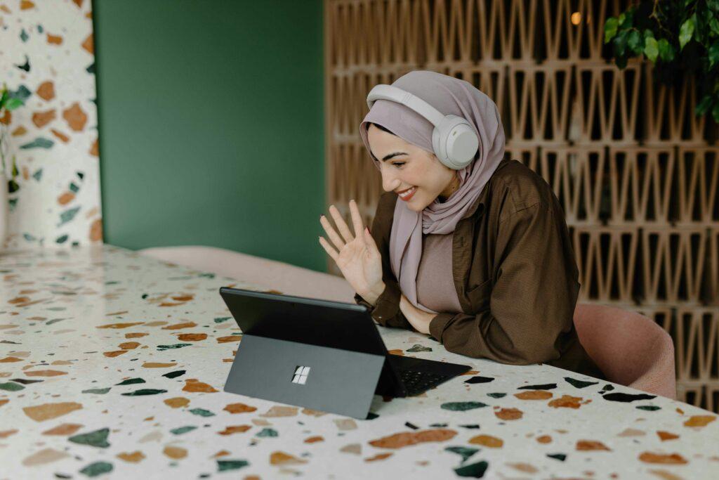 Woman in hijab and headphones on a video call using a laptop and waving.