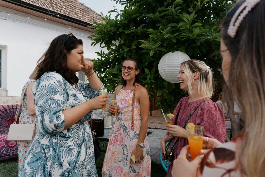 Four women are gathered outdoors, smiling and holding glasses with orange drinks. A white paper lantern hangs in the background.