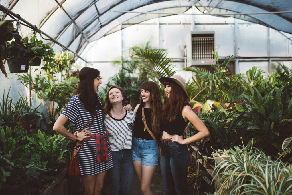 A group of four women smiling and talking closely together among rows of plants in a bright greenhouse.