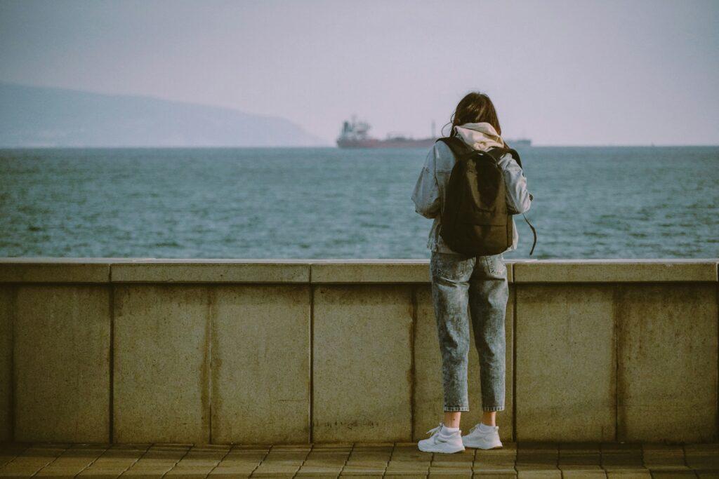 A woman wearing a denim jacket and backpack stands alone on a waterfront promenade, gazing out at a distant cargo ship on the ocean under a hazy sky.