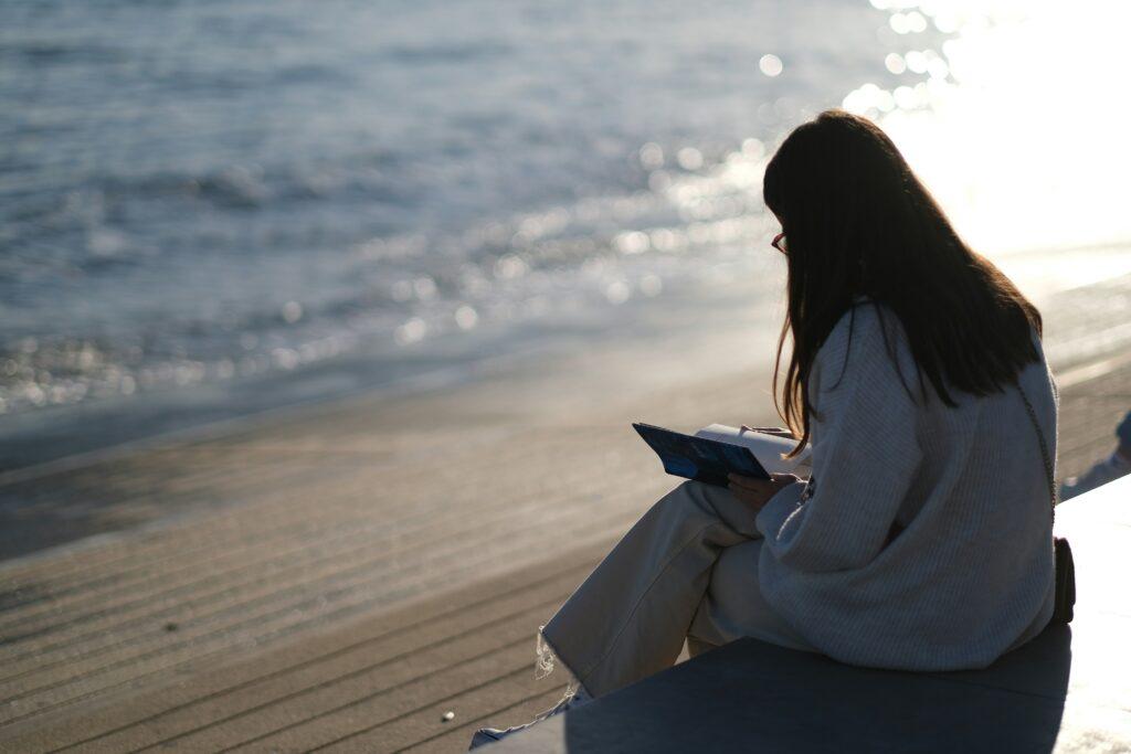 A woman with long dark hair, wearing a light sweater and cream-colored pants, sits alone by the water's edge on a sunny Summer Friday day, reading a book with the ocean gently shimmering in the background.