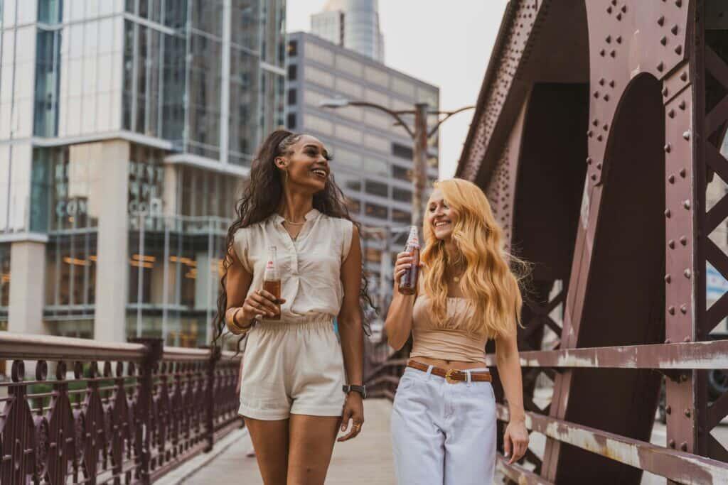 Two women walking on a city bridge, smiling and enjoying bottled drinks. One woman wears a light romper, and the other wears a strapless top with white pants. They appear relaxed and happy, with tall modern buildings in the background.