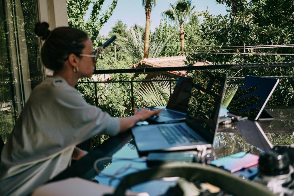Woman working remotely with multiple laptops on a balcony surrounded by lush greenery and palm trees, enjoying an outdoor work environment.