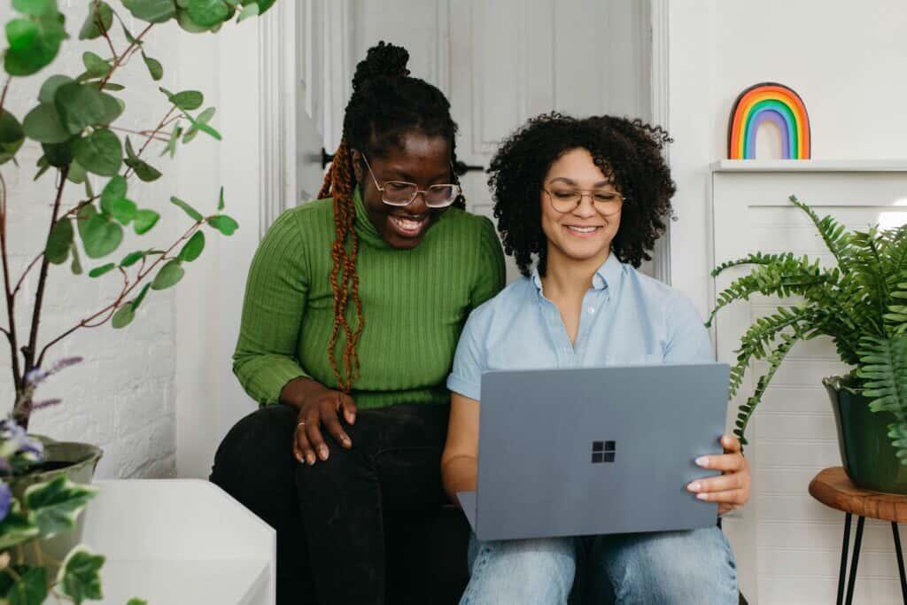 Two women smiling while working together on a laptop in a cozy indoor space decorated with plants and a rainbow ornament.