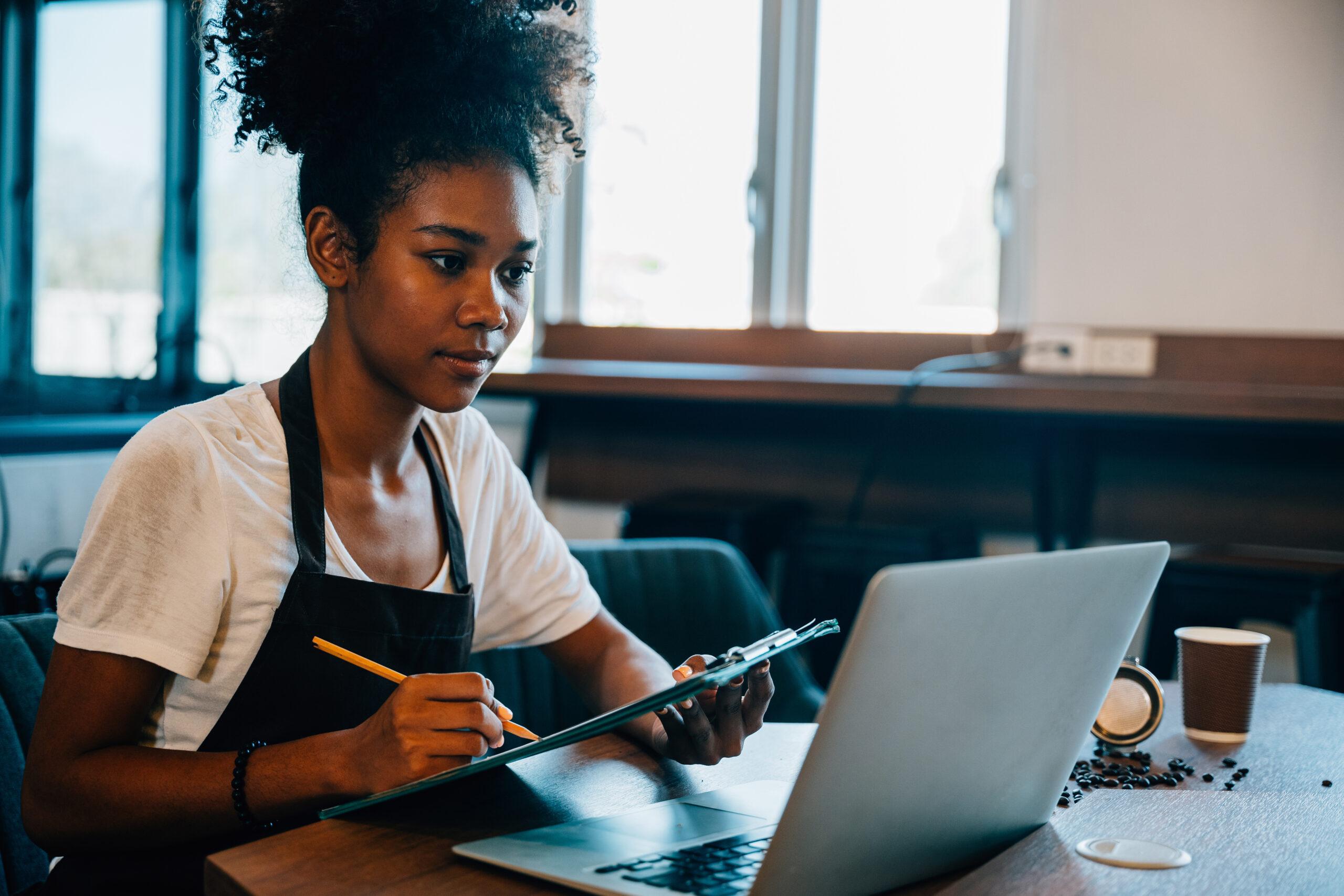 Portrait of a self-employed black woman barista at her coffee shop counter working on laptop. Confident entrepreneur managing finance bookkeeping in her own business.