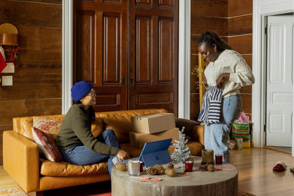 Two women share a joyful moment in a cozy, wood-paneled living room during the holidays. One woman, seated on a tan leather couch, smiles while the other stands holding a striped gift bag. Holiday decorations, a small silver Christmas tree, and gift boxes surround them.q