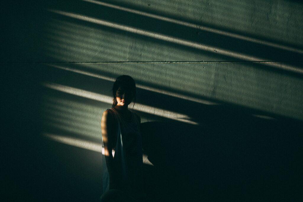 A person stands partially in shadow against a wall, with dramatic light streaming through blinds creating diagonal patterns. The mood is introspective and somber.