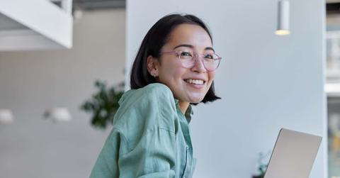 Pretty cheerful Asian woman in eyeglasses and casual clothes browses laptop computer connected to 4g internet updates software uses modern technologies poses in cafeteria looks gladfully away