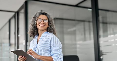 Smiling mature professional business woman holding tab looking away in office.