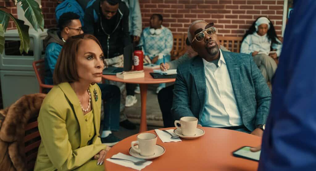 "Two adults sit at an outdoor café table with coffee cups and napkins, looking up at someone. People in casual clothes and a brick wall are in the background, giving a relaxed social setting."