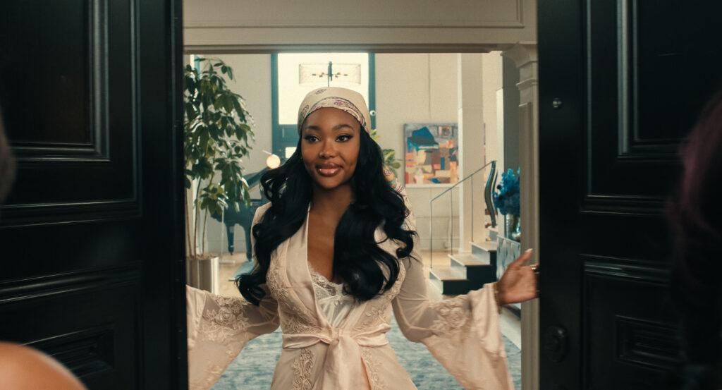 "Woman with long hair and a headband in a robe welcomes guests at a door, standing inside a modern, stylish living room with plants, art, and elegant decor visible in the background."