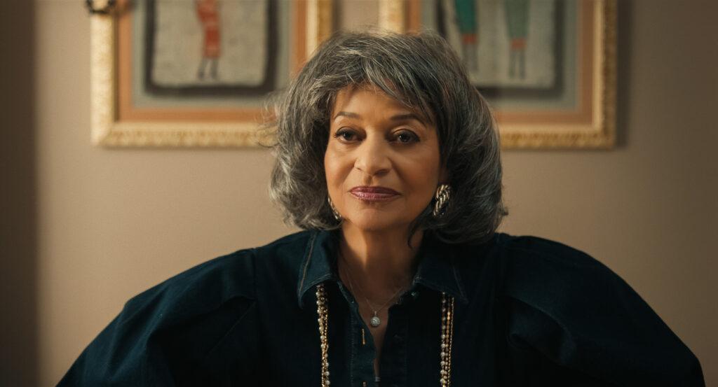 "Woman with gray hair wearing a dark blouse and gold necklace, sitting indoors in front of framed artwork on a beige wall, softly lit room."